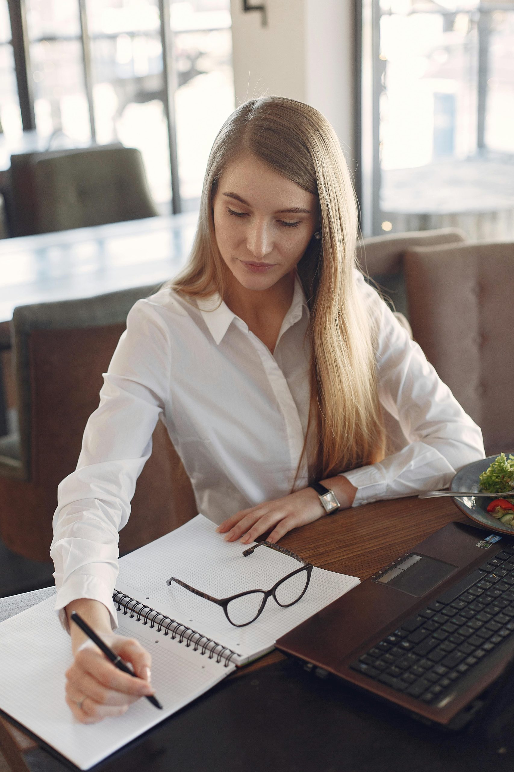 Concentrated blond businesswoman in formal wear sitting at table and making notes in copybook while working on project on laptop with fresh salad in cafe