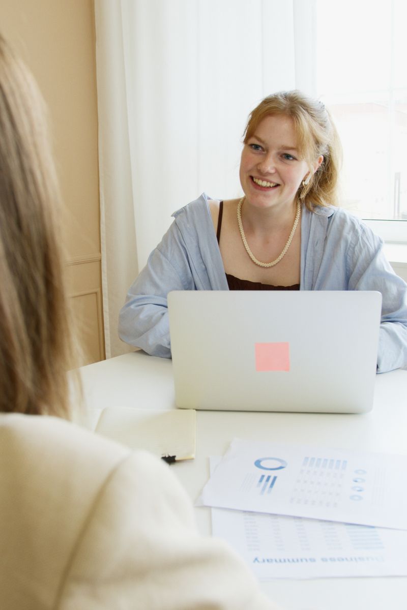 A young woman with a laptop smiling at another person in an indoor business setting.